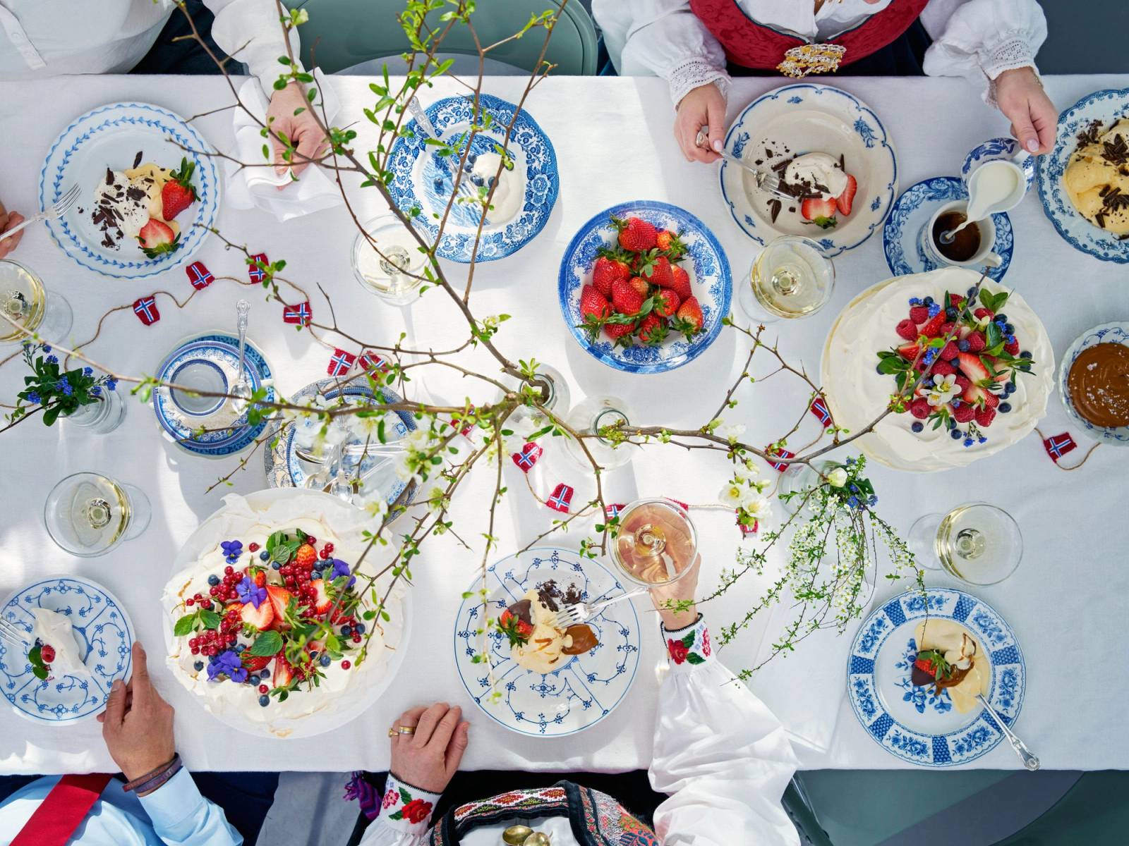 Barn og voksne feirer 17. mai med kaker og pynt på et dekket bord, med blomster og flagg i midten.