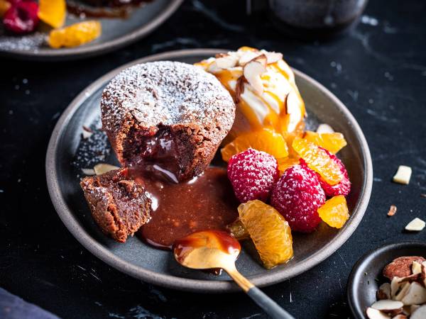 Sjokoladefondant med flytende kjerne, servert med vaniljeis, friske bær og små stykker av tørket frukt på en svart tallerken.