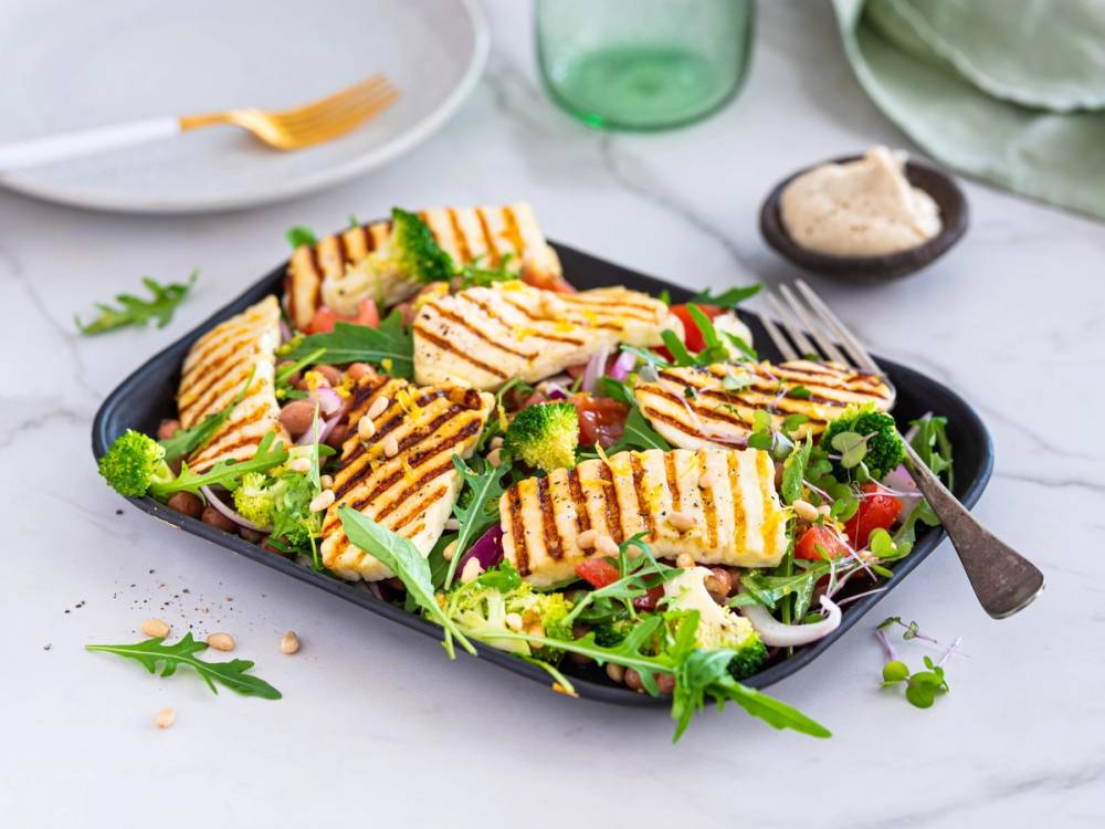 Salat med grillede halloumi-ostbiter og grønnsaker på en svart tallerken, med en liten skål med dressing eller saus ved siden av.