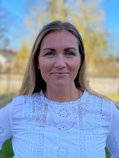 En kvinne med langt blondt hår som står utendørs med høstfarger i bakgrunnen, iført en hvit bluse med broderier.