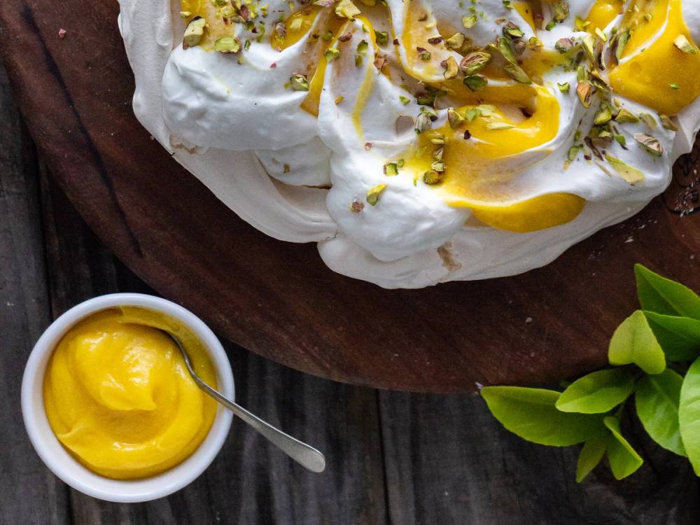 Lemon curd med sitronfarge og pyntet med sitronzest på en hvit kremet overflate, plassert på et trefat med en liten skål med mer sitroncurd ved siden av.