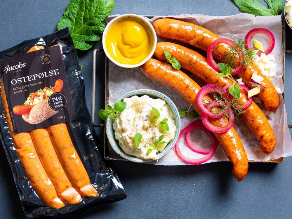 Ostepølser med sennep, løk og potetsalat på et serveringsfat, med en pakke ostepølser i forgrunnen.