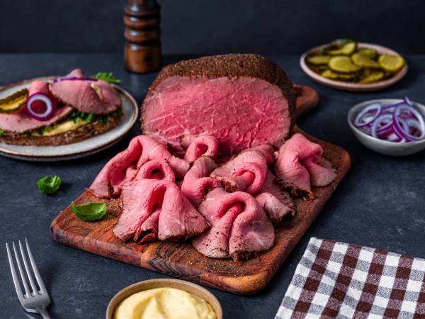 Skiver av rått storfekjøtt og kjøttdeig på en skjærebrett med tilbehør som sylteagurk og løk i bakgrunnen.