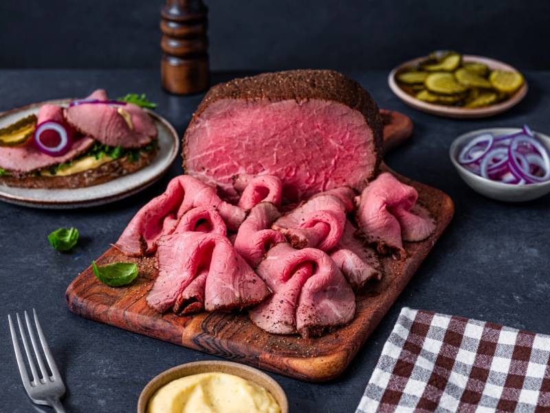 Skiver av rått storfekjøtt og kjøttdeig på en skjærebrett med tilbehør som sylteagurk og løk i bakgrunnen.