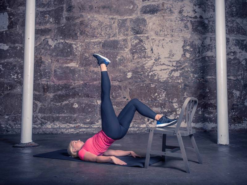 En kvinne utfører et ettbeins rompeløft med en ben opp i luften mens hun ligger på ryggen på gulvet, med en stol som støtte under det løftede beinet.