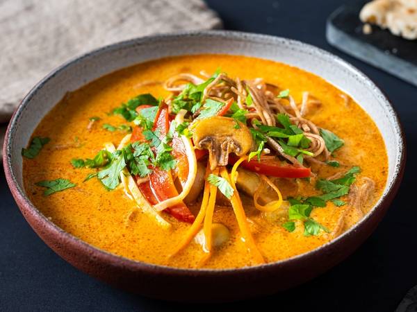 En skål med spicy grønnsaksuppe med nudler, pyntet med frisk koriander og grønnsaker, på et svart bord.