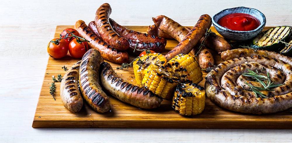 Grillpølser med grillede maiskolber og cherrytomater på et skjærebrett, med en liten skål med ketchup ved siden av.