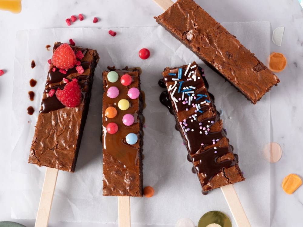 Fire hjemmelagde brownier med ulike pynt, plassert på et hvitt underlag, noen med frukt og sjokoladeknapper.