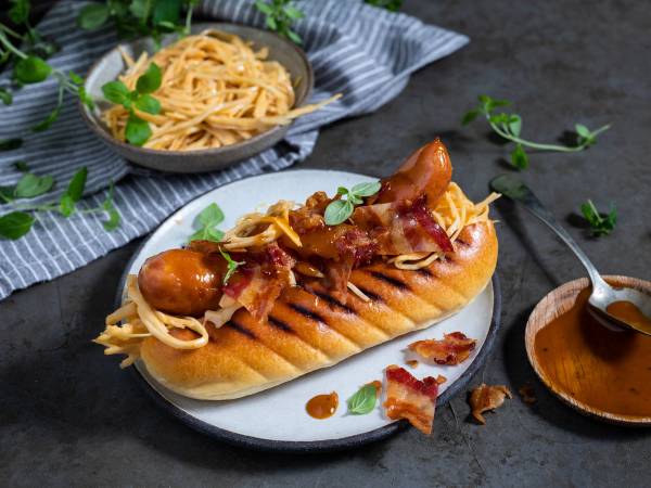 Grillet pølse i brød med bacon og løk, servert på en hvit tallerken med en skje med saus ved siden av.