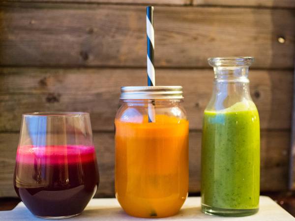 Tre ulike glass og flasker med fargerike juicer og smoothies på et bord mot en trevegg