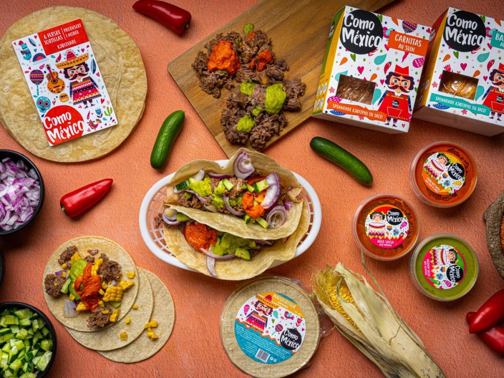 Fargerike mexicanske matvarer og produkter på et bord, inkludert tacos, små bokser med mat, og pakker med Como Mexico-produkter.