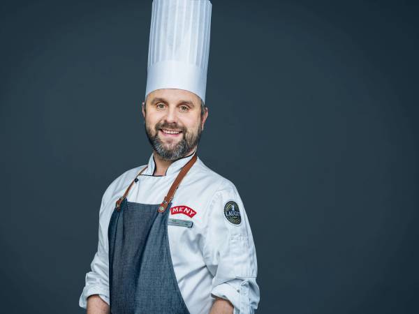 En kok i hvit uniform og kokkehatt står med armene i kors mot en mørk bakgrunn.