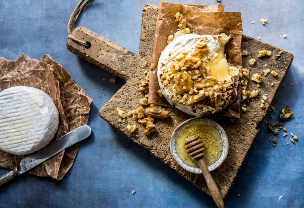 Hvitmugg på en ost med honning og nøtter på et skjærebrett, omgitt av brød og en skje med honning, på en blå bakgrunn.