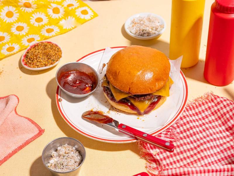 En cheeseburger med salat og tomat på en tallerken, omgitt av ketchup, sennep og salt, på et bord med en rød og hvit sjekket duk.