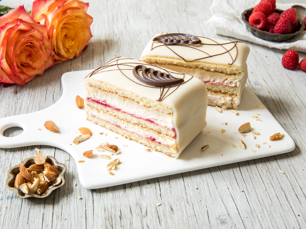 To marsipankaker med hvit sjokoladeglasur og dekorative sjokoladestykker på en hvit serveringsplate, omgitt av friske bringebær og mandler.