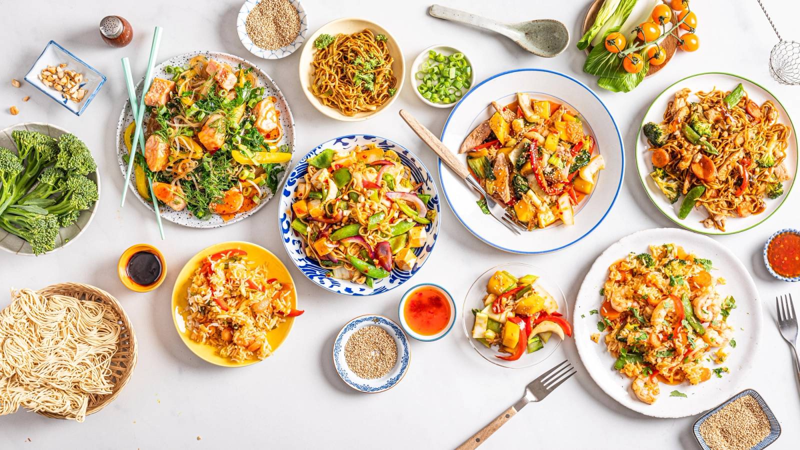 Et utvalg av wokretter og tilbehør på et hvitt bord, inkludert grønnsaker, nudler og sauser.