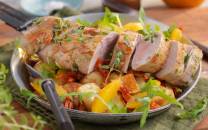 Svinefilet skåret i skiver servert på en seng av salat med sitrusbåter og urter, på en tallerken med bestikk ved siden av.