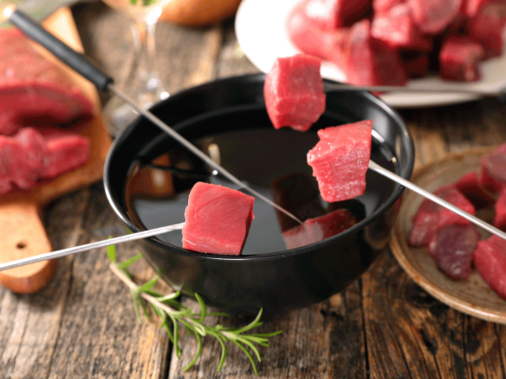 Kjøttbiter på spyd som skal dyppes i fondue, med flere kjøttstykker på tallerkener rundt.