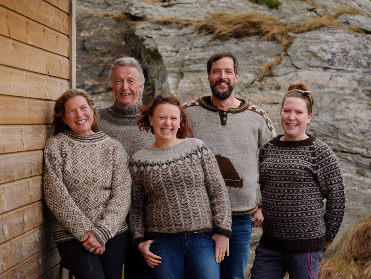 Fem personer står sammen foran en fjellvegg, alle iført strikkede gensere, og en av dem holder en havsno i hånden.