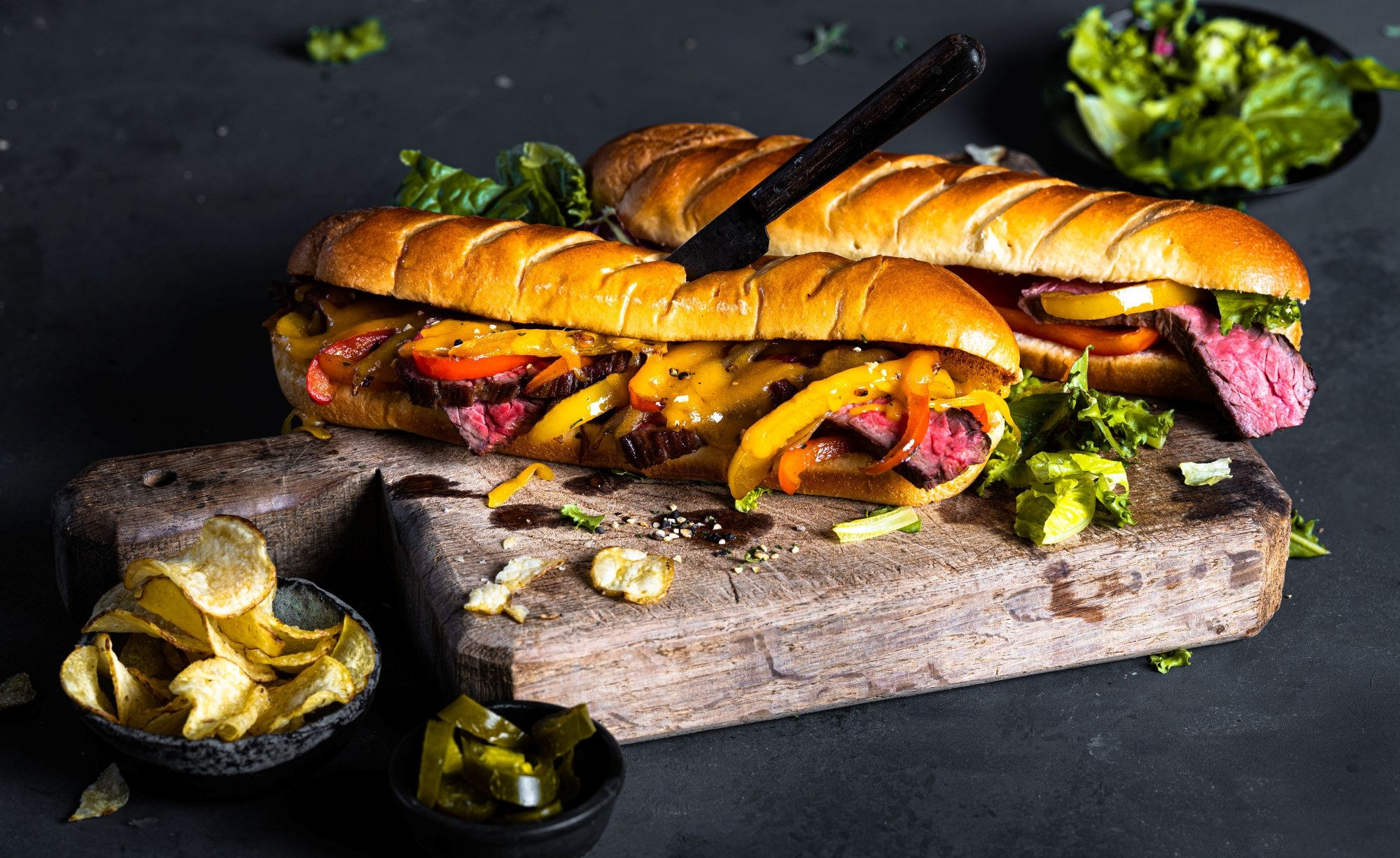 To Angus skirtsteak-sandwicher med salat og ost på en treplate, omgitt av små skåler med tilbehør.