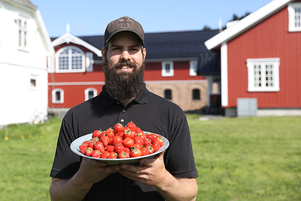 Person som holder en stor tallerken med jordbær foran røde og hvite hus i bakgrunnen på en solrik dag.
