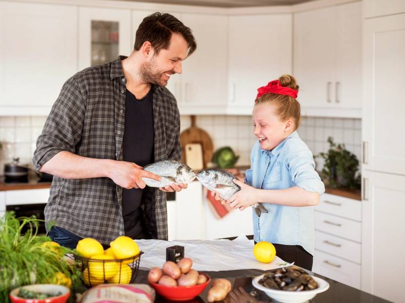 En voksen og et barn står i kjøkkenet og deler ut fisk til hverandre, med frukt og grønnsaker på benken foran dem.