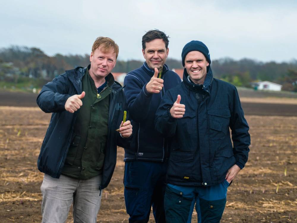 Tre personer står i en åker med åpen himmel i bakgrunnen, og de gir tommel opp mens de smiler mot kameraet.