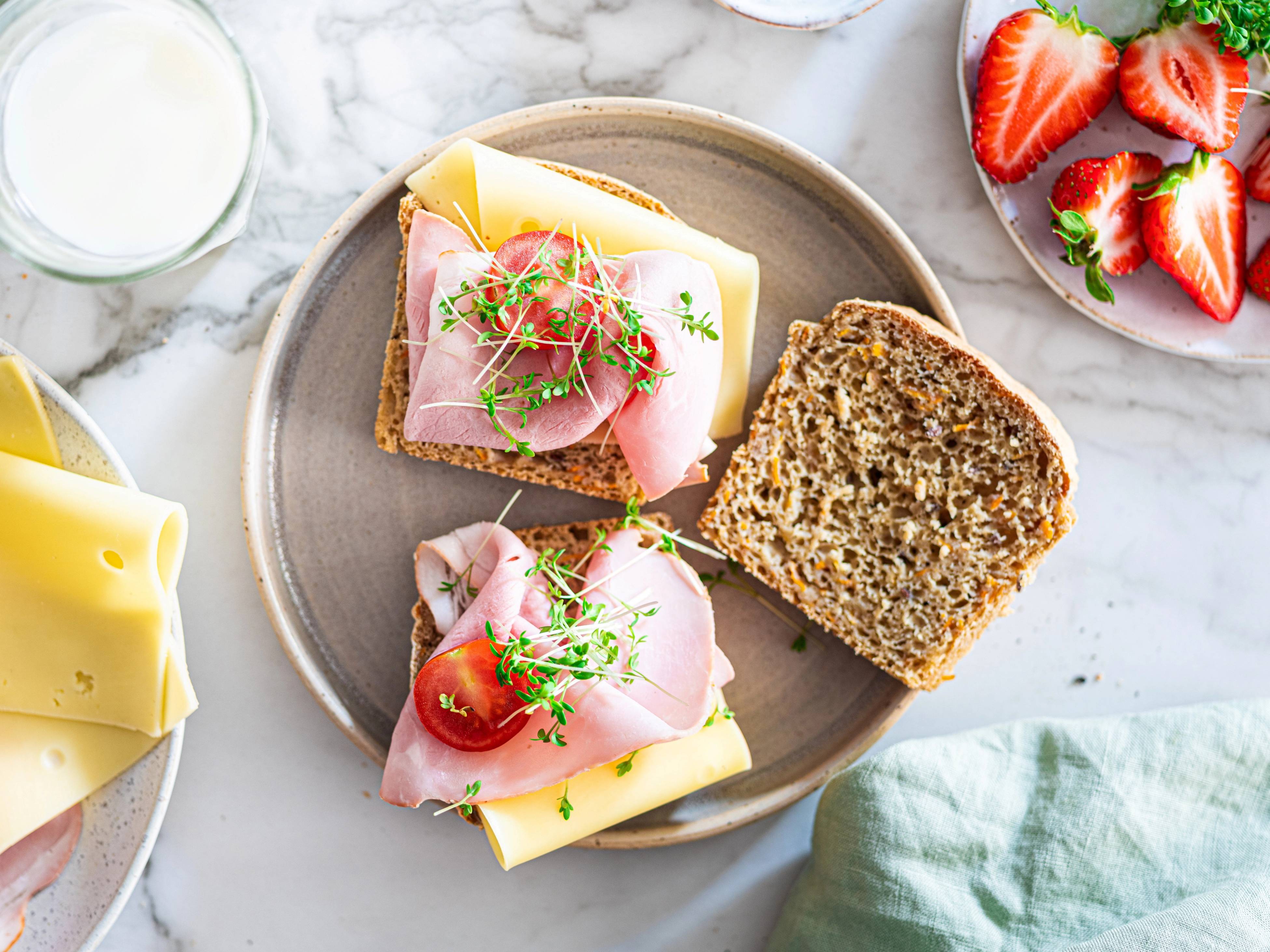 To glutenfrie matpakkebrød med pålegg som skinke, ost, tomat og spirer, servert på en tallerken med jordbær og en glass melk i bakgrunnen.