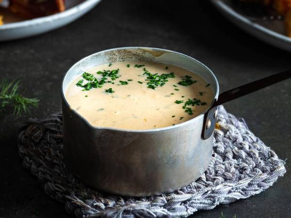 Kjele med saus til pinnekjøtt, omgitt av tallerkener med kjøtt og kålrotstappe på en mørk bakgrunn.