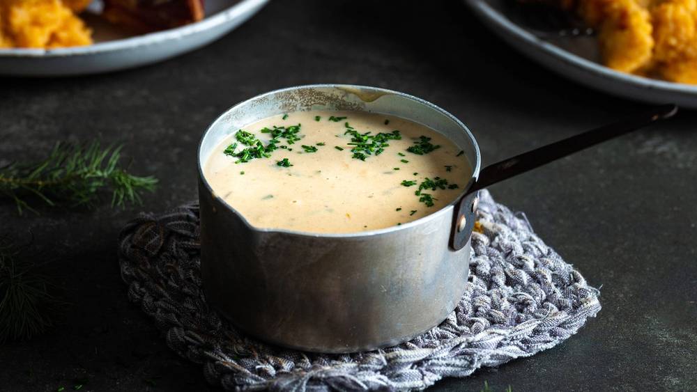 Kjele med saus til pinnekjøtt, omgitt av tallerkener med kjøtt og kålrotstappe på en mørk bakgrunn.