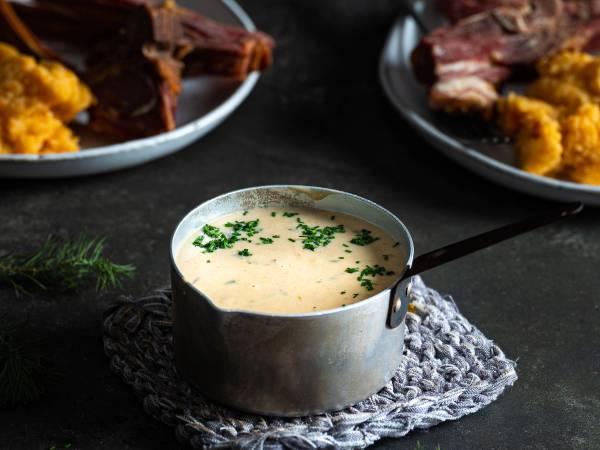 Kjele med saus til pinnekjøtt, omgitt av tallerkener med kjøtt og kålrotstappe på en mørk bakgrunn.