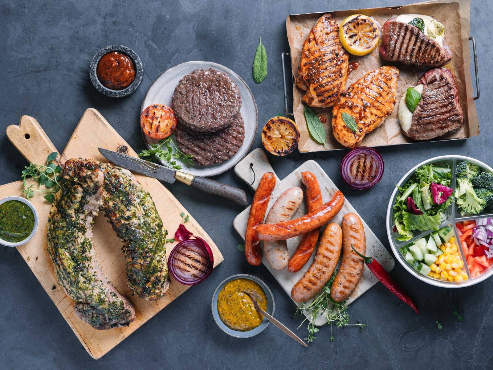 Grillmat med pølser, kylling, biff og tilbehør som salat, grønnsaker og saus på et utendørs dekket bord.