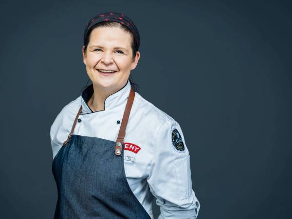 En person iført kokkeuniform og forkle, med en mørk bakgrunn, uten å vise ansiktet tydelig.