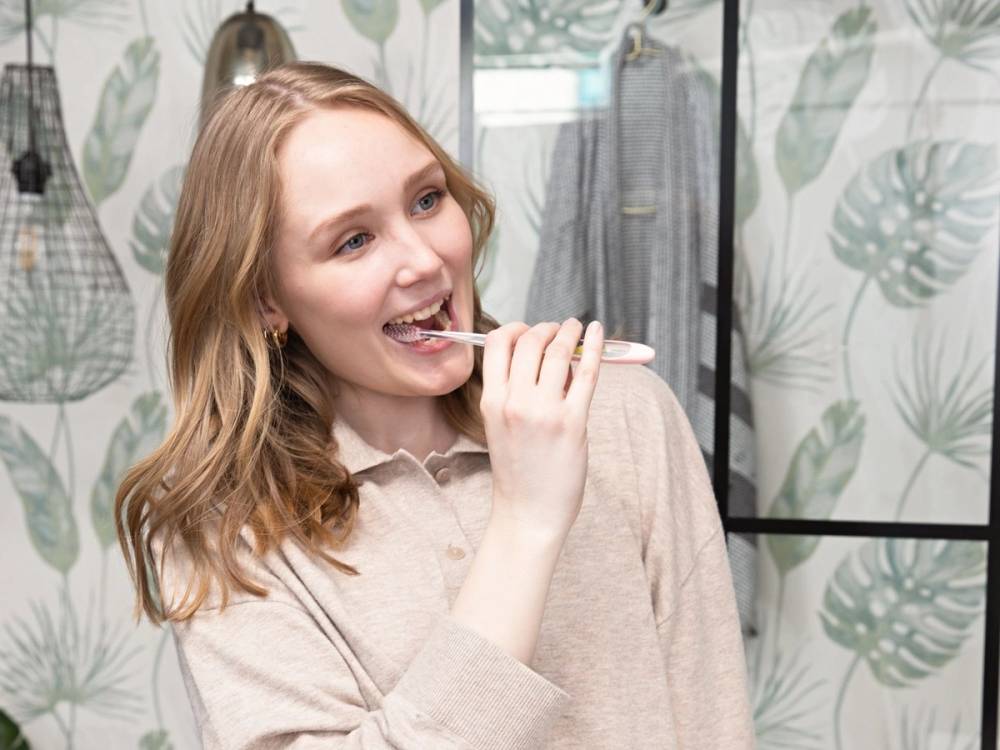 En person med langt, blondt hår tar et speilbilde i en butikk med grønne plante- og bladmønstrede veggdekorasjoner i bakgrunnen.