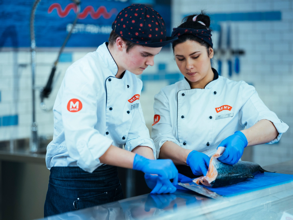 To ansatte i kokkeklær og blå hansker som forbereder fisk i et kjøkkenmiljø.