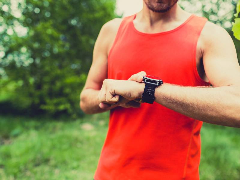 En mann i rødt treningsskjorte ser på klokken på håndleddet sitt under utendørs trening, med grønt løv og trær i bakgrunnen.