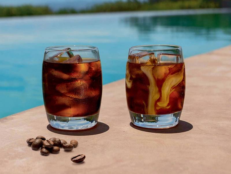 To glass med kald kaffe med is, plassert på et bord ved en svømmebassengkant, med kaffebønner foran.