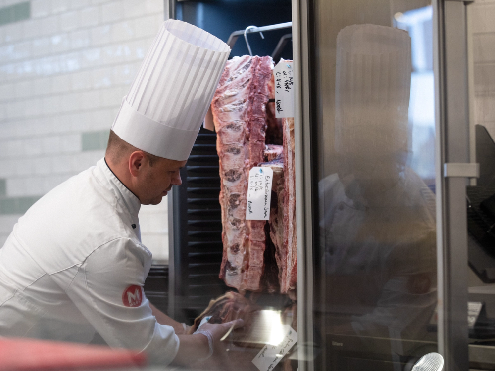 En kok i hvit kokkeuniform og lue inspiserer tørket oksekjøtt på et kjølerom.