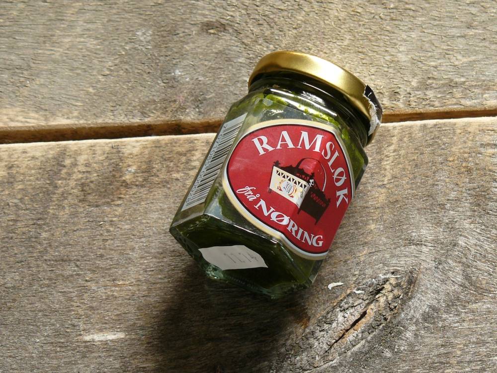 Et glass med Ramsløk-urte på et trebord med en gullfarget lokk og etikett som sier 'Ramsløk' og 'Tørking'.