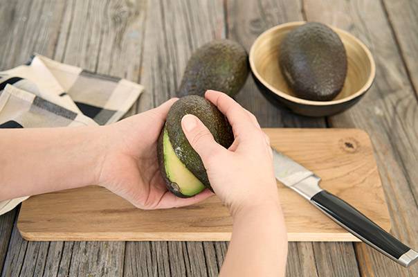 Person skreller en avocado over et skjærebrett med to avocados i en skål i bakgrunnen.