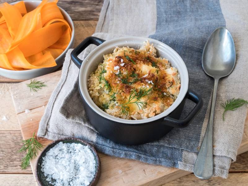 Hjemmelaget fiskgrateng i en liten gryte med dill og ost på toppen, servert på et bord med en skje og en skål med salt ved siden av.