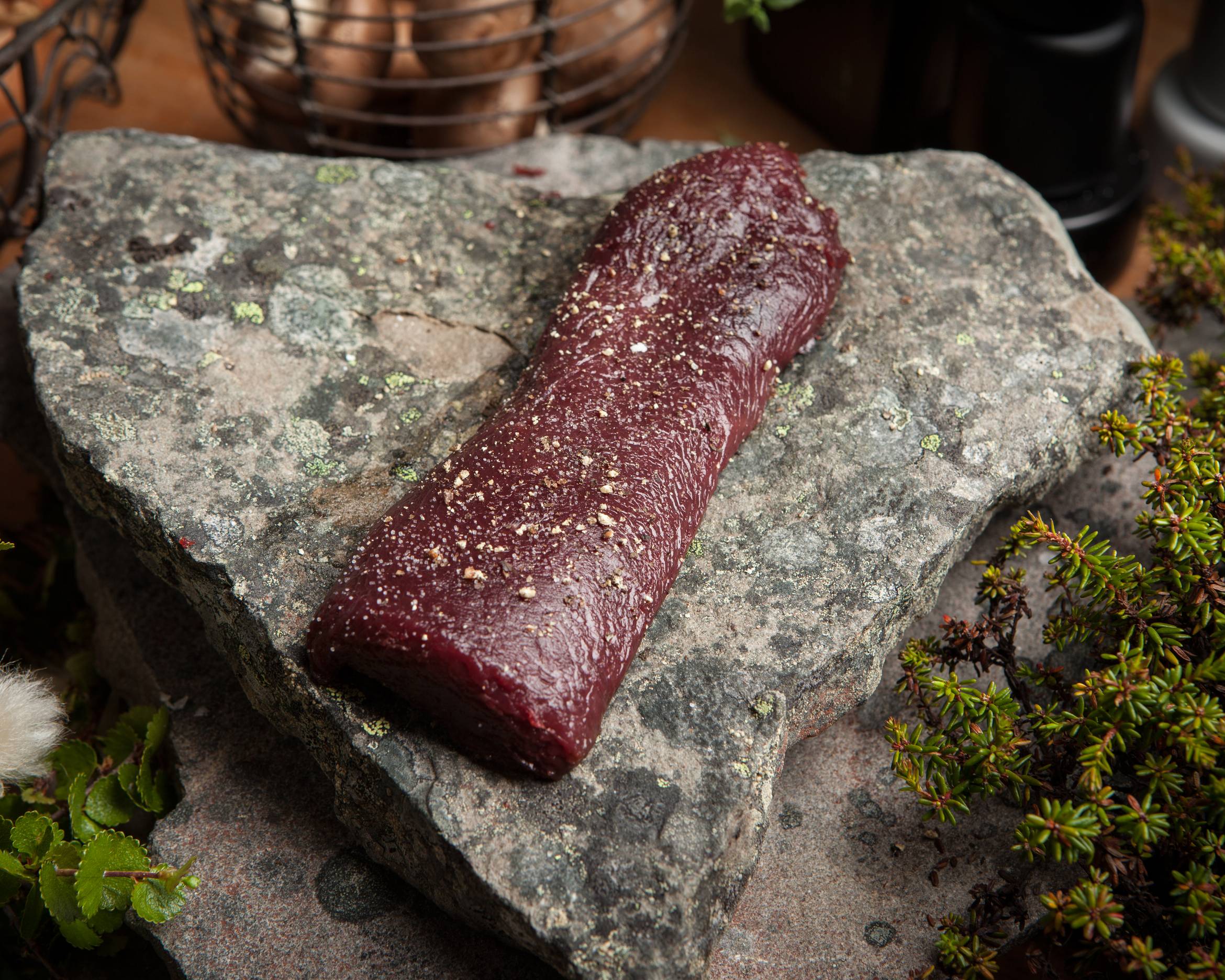 En rå ytrefilet av rein ligger på en stor stein, omgitt av små planter og urter.