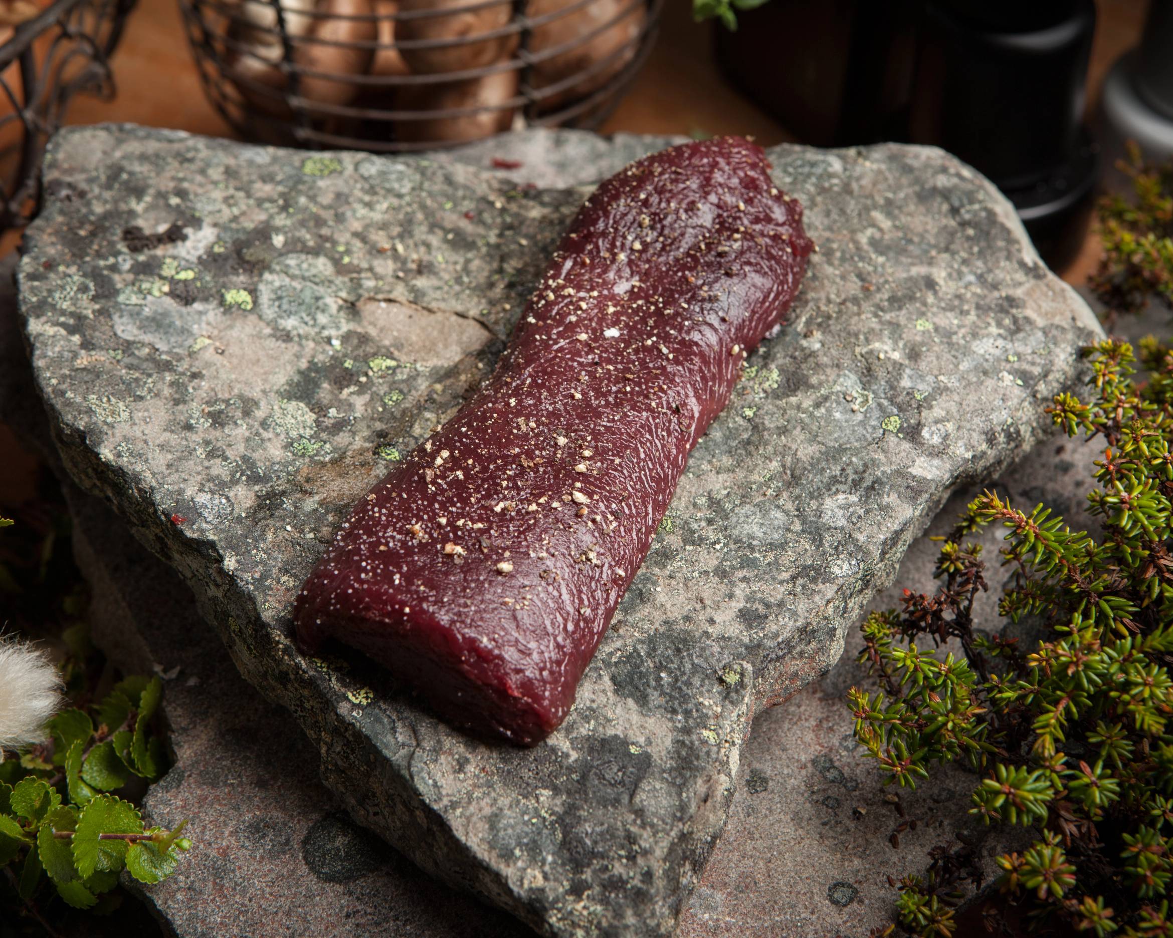 En rå ytrefilet av rein ligger på en stor stein, omgitt av små planter og urter.