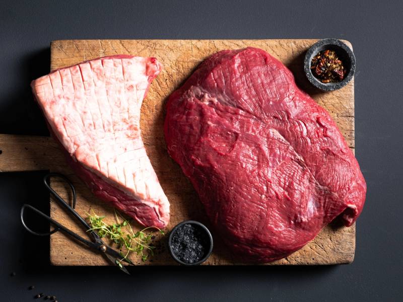 Et stykke angus tritip kjøtt og en morbrad på et skjærebrett med krydder og urter.