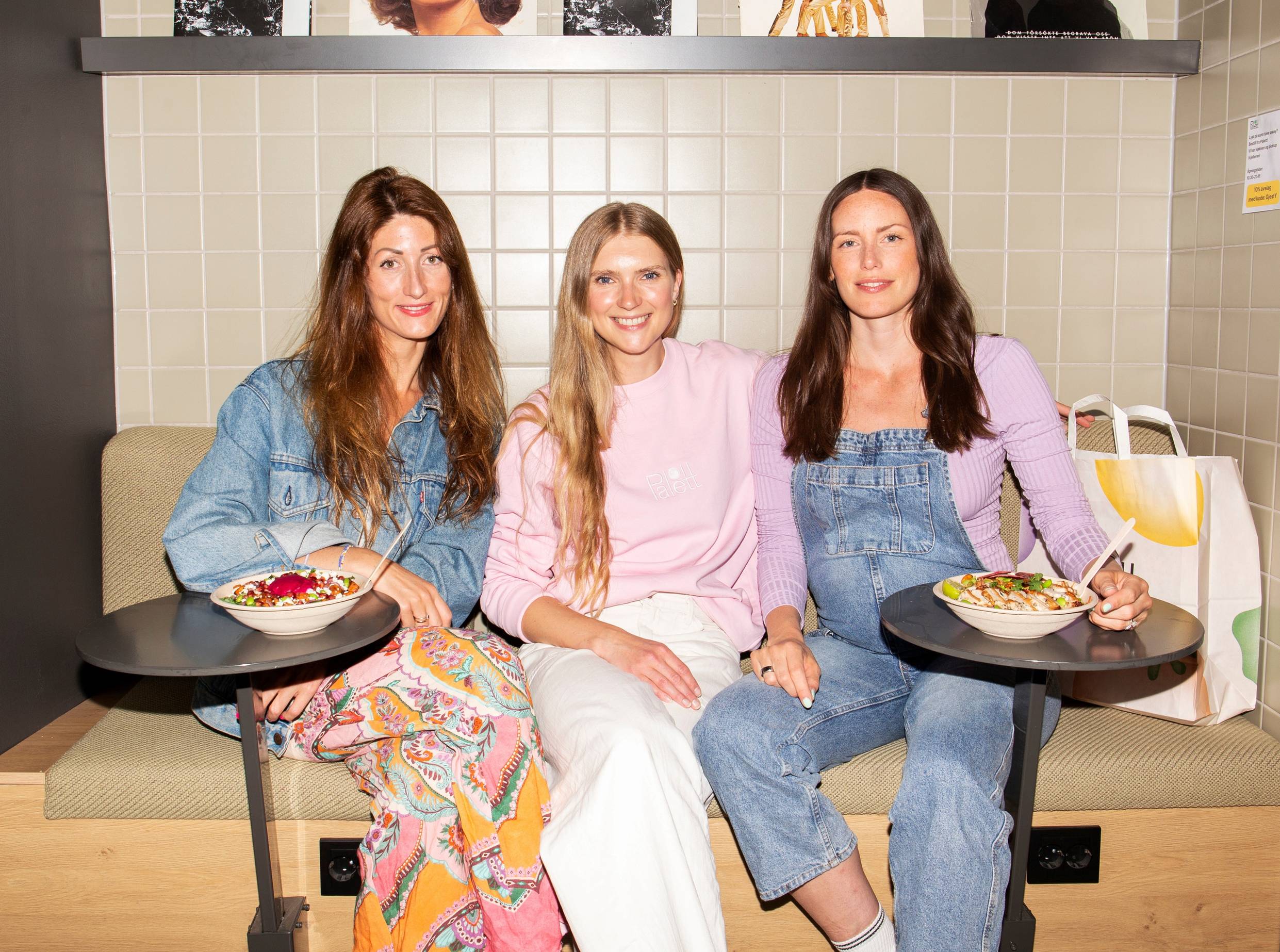 Tre kvinner sitter på en kafé, to med mat på tallerkener foran seg og en med en kopp, i en moderne innendørs setting.
