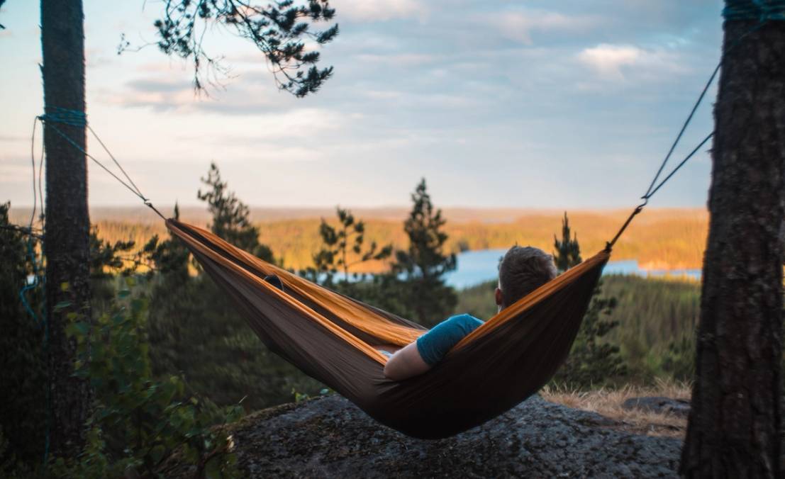 En person ligger i en hengematte mellom to trær med utsikt over en innsjø og skog i bakgrunnen, under en klar himmel.