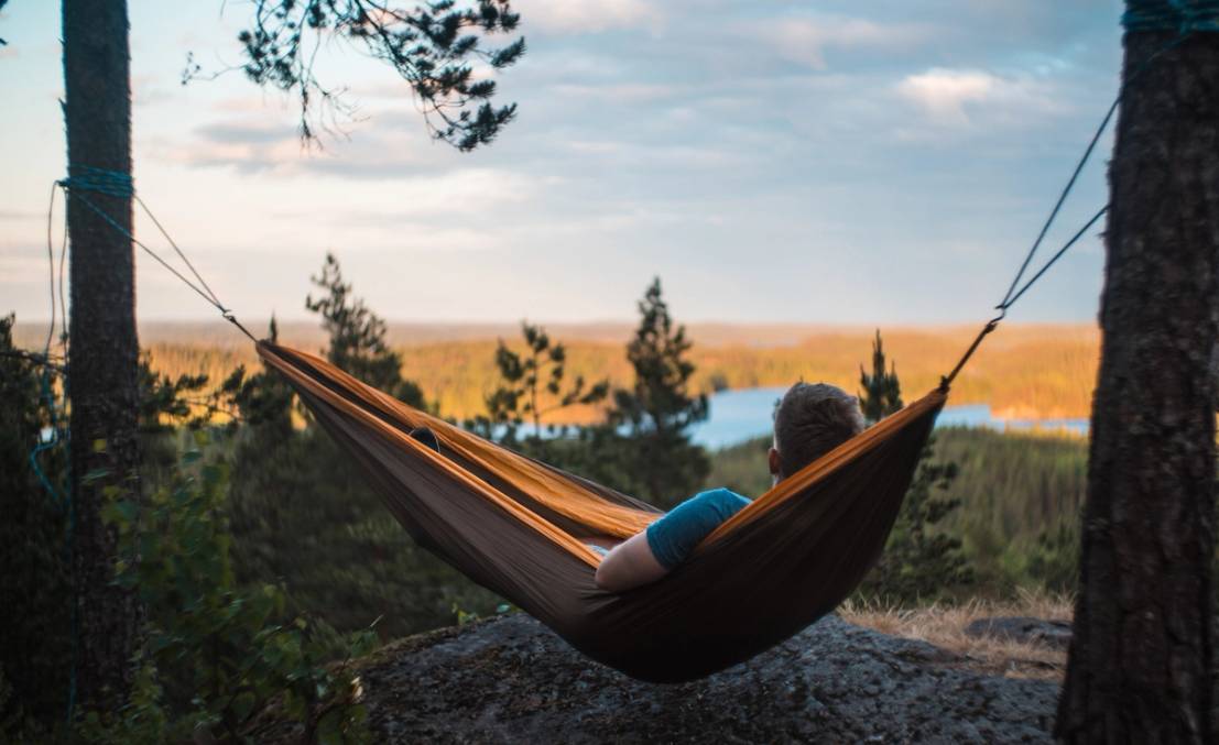 En person ligger i en hengematte mellom to trær med utsikt over en innsjø og skog i bakgrunnen, under en klar himmel.