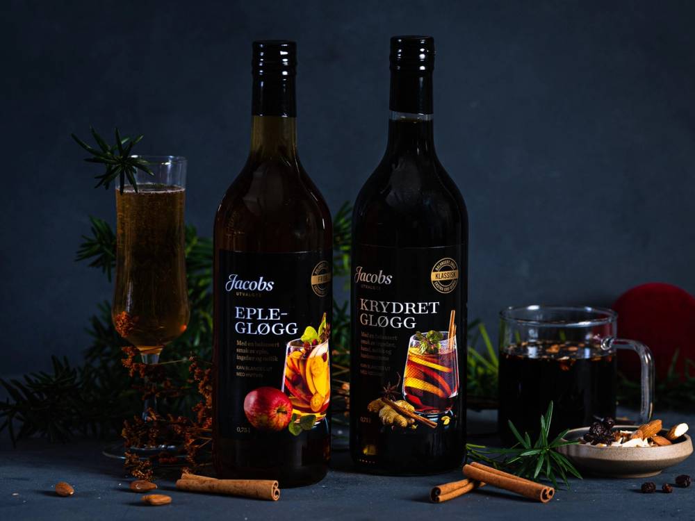 To flasker med Jacobs utvalgte gløgg, en med eplegløgg og en med krydret gløgg, omgitt av kanelstenger, nellik og granbar på et mørkt bord.