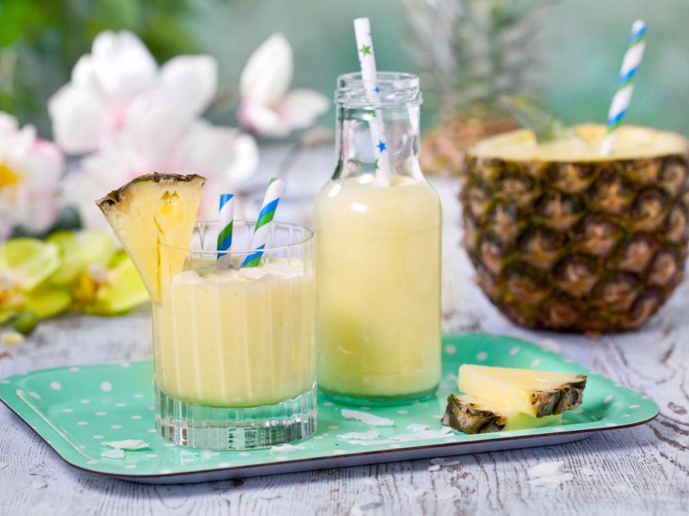 En tropisk smoothie servert i et glass med en skive ananas og et sugerør, med en hel ananas i bakgrunnen.