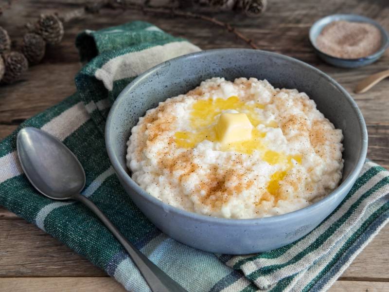 Risgrot med smør og sukker servert i en blå bolle på et bord med skje og strikket klut, omgitt av små skåler med tilbehør.